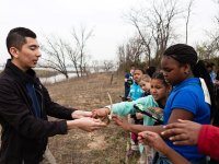 A group of young students are standing in a line on dead grass. It's overcast. There are bare-branched trees and a river nearby. They're holding black and green tablets. Their male teacher is holding antlers, letting the students take turns touching it.