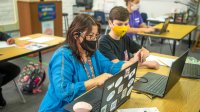 Teacher helps student working on Chromebook while wearing masks