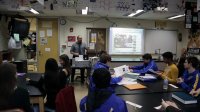 A classroom full with students and a teacher. Teacher is teaching the students with the presentation in front of the classroom.