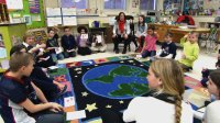 Students and teachers sitting on the classroom floor talking to each other.