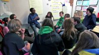 A student is speaking in front of the classroom among his friends listening to him.