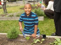A young boy is kneeling on the grass beside a garden bed with a shovel in his hand, smiling at the camera. 