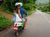 A man and woman on a motorcycle are driving on a road with tropical trees surrounding them on both sides.
