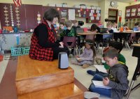A teacher working with elementary students, who are writing in notebooks near a smart speaker device in a classroom