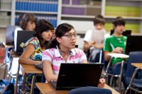 High school students sit at their desks with laptops open and attention on their teacher