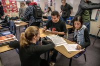 Teacher kneeling down working with two high school students sitting at their desks