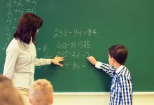 Elementary school teacher works with student on math problem on chalk board in classroom