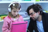 Jordan Shapiro sitting with an elementary student who is using a tablet and wearing headphones