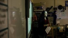 A teacher standing at the front of class, her face illuminated by a projection on the whiteboard behind her