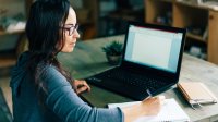 Woman writing in notebook in front of laptop at home