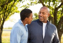 Standing outside between two trees, a father and son are looking at each other, smiling, and hugging each other with one arm. 