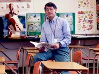 Man sitting on a desk in a biology classroom holding an open book
