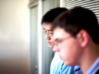 A closeup of two young, adult males looking dejected with their heads down. They're standing against a window with closed blinds. 