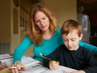 A mom is helping her young boy with homework in the living room of their house. They're both sitting at a table with a text book and workbook opened up.
