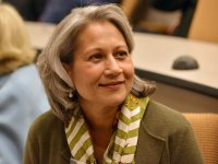 A close up of a woman from the chest up sitting in a lecture hall. She's smiling, looking off to the side, wearing a green sweater and a striped white and green scarf around her neck. She has layered, shoulder-length gray hair, peppered with brown.