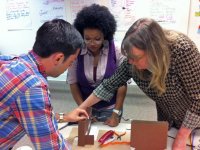 One male and two female teachers are leaning over a classroom table, building something together. They have two pieces of cardboard cut, standing up like mini buildings, one with an open door, and a piece of wire is connecting them.