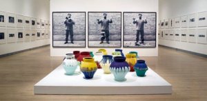 Three full length pictures behind a display of 15 colorful pots of Ai Weiwei holding, dropping an urn, and seeing the pieces on the ground
