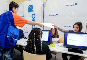 Girls at a table with computers and a boy standing handing paperwork to one of the girls