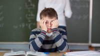 A middle school male student sits at his desk, frowning and holding his head in his hands. 