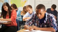 A class of high school students are writing in notebooks at their desks. 