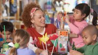 A teacher is smiling, sitting at a classroom table with four young kids. They're building things out of pipe cleaners, Kleenex boxes, and toilet paper roles.