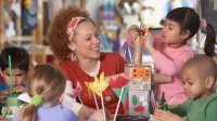 A teacher is smiling, sitting at a classroom table with four young kids. They're building things out of pipe cleaners, Kleenex boxes, and toilet paper roles. 