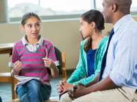 Two young girls around middle school age and a male teacher are sitting in chairs set up in a circle on the carpet in their classroom. The teacher and one of the girls is listening to the other girl speak.