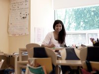 A teacher is sitting by herself in her classroom by a window. At her desk, with a pencil in hand and paper in front of her, she's smiling at the camera. 