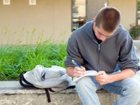 Older boy sitting on a large cement planter in a school courtyard writing in a spiral notebook