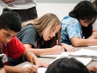 Students are hunched over their desks, writing in notebooks, with their teacher looking upon them.