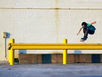 A young teenage boy in a teal t-shirt and brown pants is in the air on a skateboard. Both of his arms are extended out to his sides and his knees are up to his chest. He's hovering over a yellow beam against an off-white brick wall.