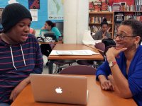A high school student is sitting across a table from an adult in a library, and they're talking to each other. There are bookshelves against the wall behind them, as well as another table, and students sitting at desks.