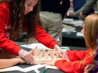 Two students are sitting, and one student is standing, all their hands are touching a popsicle stick bridge that they're gluing together.