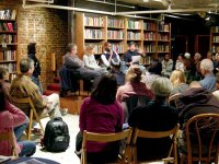 Room of seated adults surrounded by book shelves with four people front and center speaking