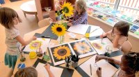 Five first grade girls sit at a round table painting portraits of sunflowers.