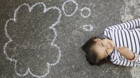 A smiling six-year-old girl lies on the school playground with a thought bubble drawn in chalk above her head.