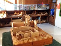 In the center of the classroom there is a big block house on top of a wooden block on top of a green table. Against one wall are shelves filled with blocks.