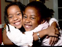 Two young girls are smiling and hugging each other, looking at the camera.