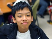 A closeup of a young boy in his preteens sitting in class. He's looking directly at the camera with a close-mouthed smile. The rest of the photo is out of focus. 