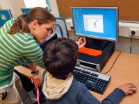 A teaching is standing next to a sitting student, talking to him and pointing at the desktop computer screen in front of him.