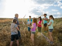 Nine students are standing in a vast field with a male teacher among tall, dry grass. They're placing hand-made windmills in the field.