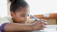 A young girl sits at her desk, writing with a pencil.