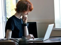 A young red-headed boy is sitting at his desk looking at his laptop, his chin resting on one hand and his other hand on the laptop keyboard. 