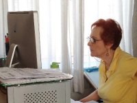 An older, red-headed woman with glasses and a yellow top is sitting in an office, looking at a computer screen.