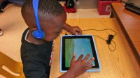 A bird eye's view of a young boy sitting at his classroom desk with blue headphones on, playing a game on a blue tablet.