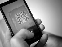 A black and white closeup of a hand holding a cell phone with a QR code on the screen. 