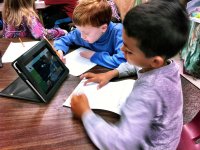 Two young boys are sitting at a wooden, circular table in a classroom. They're sharing a tablet, propped up, watching something on the screen, and filling out a worksheet.