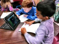 Two young boys are sitting at a wooden, circular table in a classroom. They're sharing a tablet, propped up, watching something on the screen, and filling out a worksheet. 