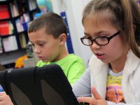 A young girl wearing glasses, pigtails, and a white vest over a gray, long sleeve shirt is sitting, looking at a tablet that's propped up on the table. She is sitting beside a boy in a bright green shirt who's also looking at a tablet.