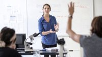 A teacher in a science classroom talks as a student raises their hand.